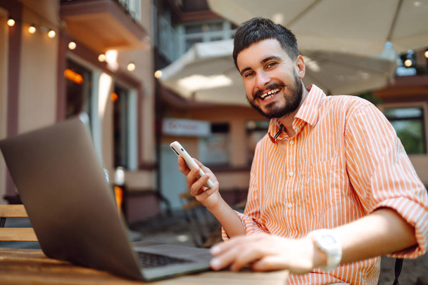 Smiling young man working in a cafe on the street with a laptop and phone. Freelance business concept. Business, blogging, freelancing, education concept. Modern lifestyle. - Foto, imagen