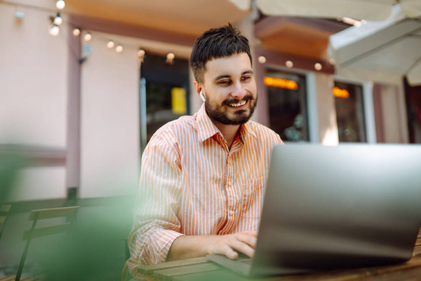 Smiling young man working in a cafe on the street with a laptop and phone. Freelance business concept. Business, blogging, freelancing, education concept. Modern lifestyle. - 写真・画像