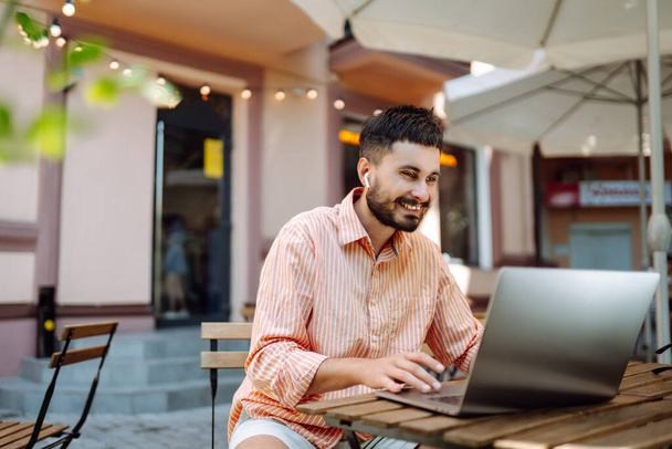 Smiling young man working in a cafe on the street with a laptop and phone. Freelance business concept. Business, blogging, freelancing, education concept. Modern lifestyle. - Fotoğraf, Görsel