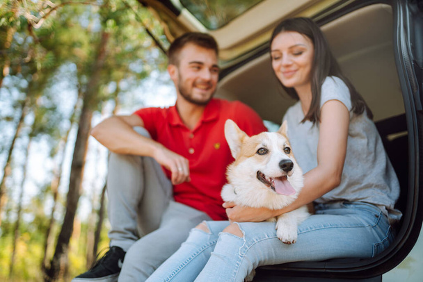Lovely young couple with a dog sits in the back seat of a car. Happy man with a woman and his beloved dog travel together. Concept of active lifestyle, travel, weekend, relax. - Photo, Image