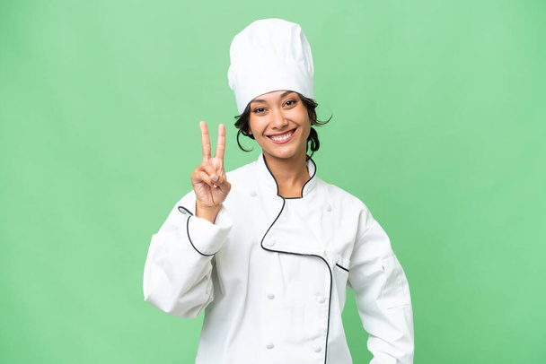Jonge chef-kok Argentijnse vrouw over geïsoleerde achtergrond glimlachen en tonen overwinning teken - Foto, afbeelding