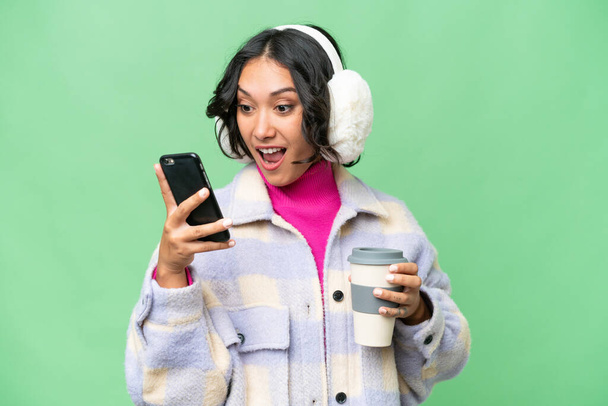 Jonge Argentijnse vrouw draagt wintermuffs over geïsoleerde achtergrond met koffie om mee te nemen en een mobiele - Foto, afbeelding