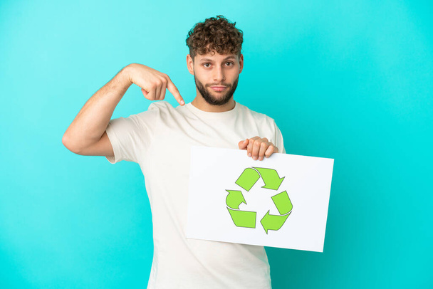 Young handsome caucasian man isolated on blue background holding a placard with recycle icon and  pointing it - Foto, imagen