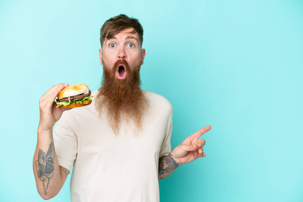Roodharige man met lange baard houdt een hamburger geïsoleerd op blauwe achtergrond verrast en wijzend kant - Foto, afbeelding