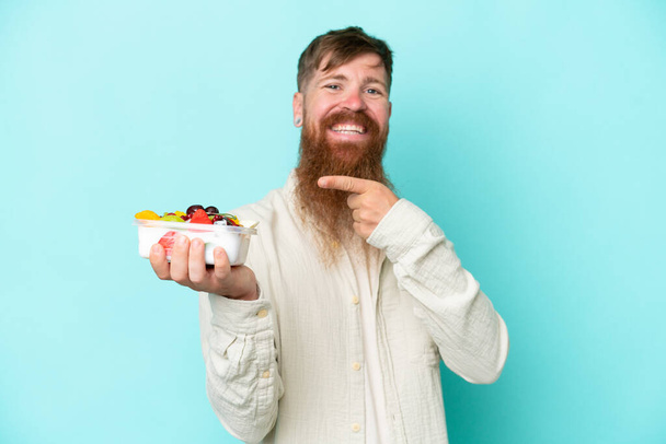 Rotschopf mit langem Bart hält eine Schale mit Früchten isoliert auf blauem Hintergrund und zeigt darauf - Foto, Bild