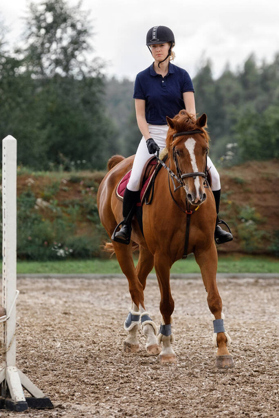 Ένα κορίτσι με τη μορφή αναβάτη ιππεύει ένα άλογο στον ιππόδρομο. Μαθήματα ιππασίας, ιππασία. - Φωτογραφία, εικόνα