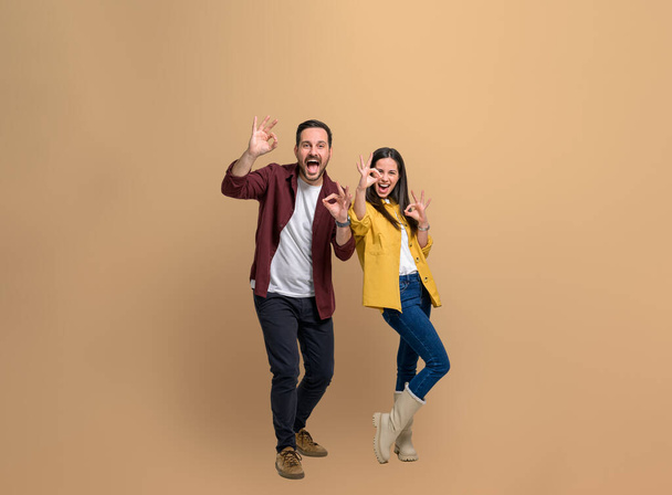 Full length of stylish young couple showing OK Okay signs and screaming ecstatically while looking at camera. Excited man and woman gesturing while posing happily over beige background - Фото, изображение