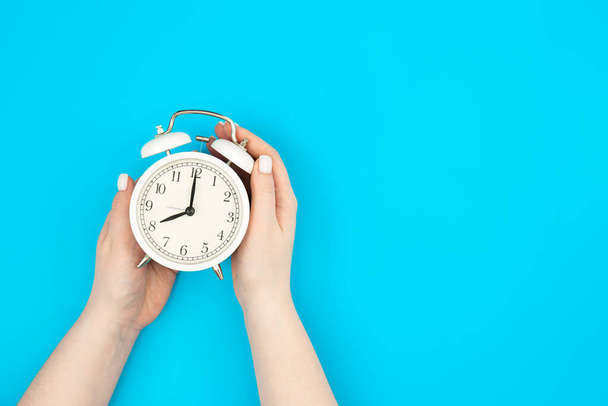 Reloj despertador blanco en manos femeninas sobre fondo azul, vista superior, concepto de gestión del tiempo, fecha límite y trabajo en la oficina. - Foto, imagen