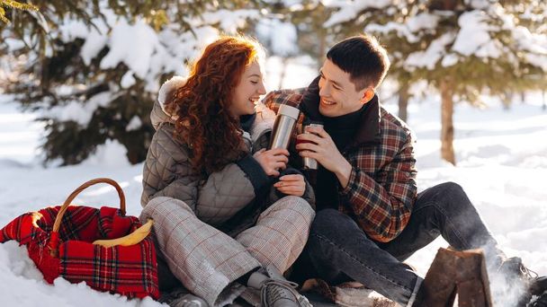 Young people have a picnic in the park on a sunny winter day. Young people sitting in a snowy park drink hot tea from a thermos and warm themselves near the fire. High quality photo - Φωτογραφία, εικόνα