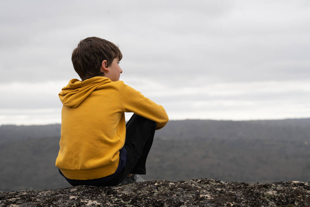 Спокойный мальчик сидит на вершине горы и смотрит на горизонт с копировальным пространством.. - Фото, изображение