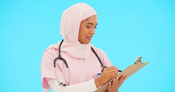 Woman, doctor and writing on clipboard in studio for information or medical record. Islamic or muslim female and professional nurse in hijab with list for healthcare insurance on blue background. - 写真・画像