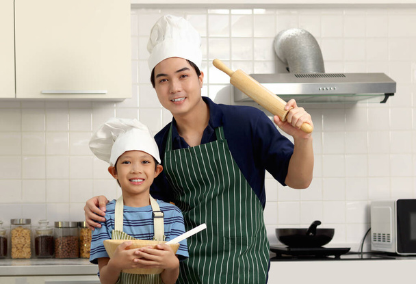Portrait of happy Asian father and son kid with apron chef hat hold wooden rolling kneading stick and bread flour bowl at kitchen, dad present and boy child make bread together, family chef cooking. - Photo, Image
