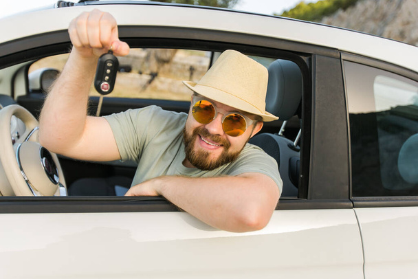 Car driver man smiling showing new car keys and car. Bearded guy driving rented cabrio in summer vacation - Fotó, kép
