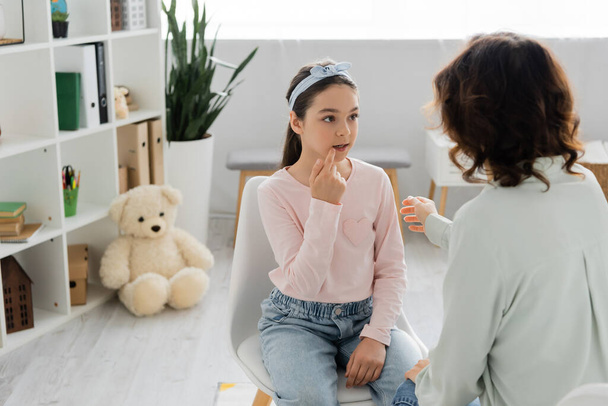 Preteen girl talking and pointing at mouth near blurred speech therapist in consulting room  - 写真・画像