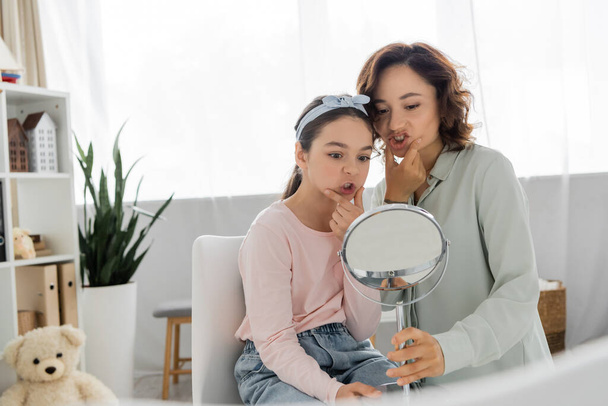 Pupil touching cheeks and looking at mirror near speech therapist in consulting room  - Photo, Image