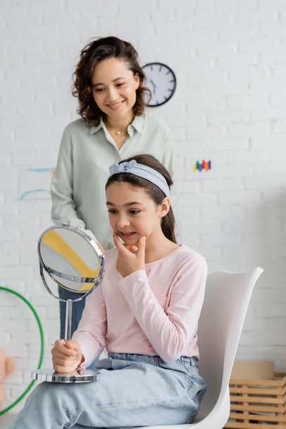 Preteen child holding mirror and speaking near smiling speech therapist in consulting room  - Photo, Image