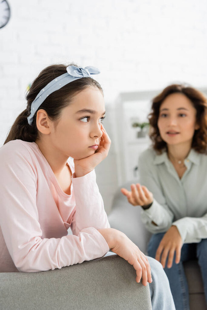 Pensive preteen child sitting near blurred psychologist in consulting room  - Фото, изображение