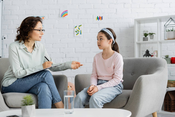 Psychologist writing on clipboard and talking to preteen girl in consulting room  - Zdjęcie, obraz