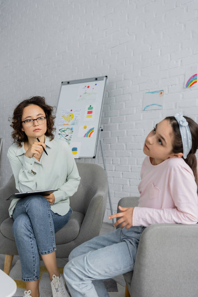Focused psychologist holding clipboard and looking at pensive child in consulting room  - Photo, image