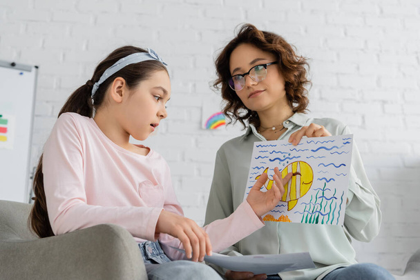 Chica preadolescente hablando y apuntando a dibujar cerca de psicólogo en la sala de consulta  - Foto, imagen