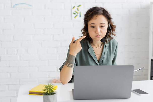 Speech therapist in headset pointing at mouth and holding clipboard during video call on laptop in consulting room  - 写真・画像