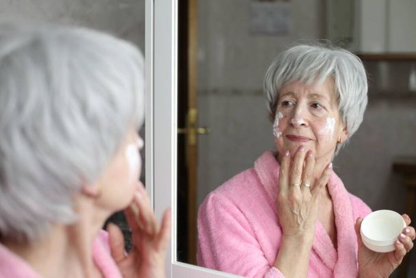 close-up portret van volwassen vrouw aanbrengen gezichtscrème in de voorkant van de spiegel in de badkamer - Foto, afbeelding