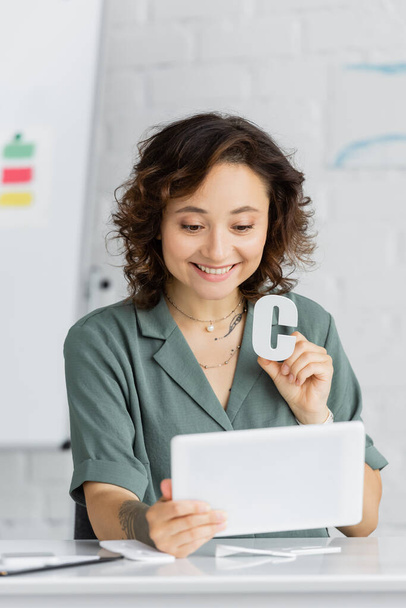 Позитивный логопед держит письмо и цифровой планшет во время онлайн-урока в консультационном кабинете  - Фото, изображение