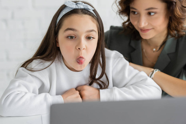 Preadolescente chica sobresaliendo lengua durante la lección de terapia del habla en el ordenador portátil cerca borrosa mamá en casa  - Foto, Imagen