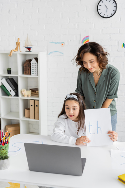Smiling mother standing near daughter holding paper with letters during speech therapy online lesson at home  - Foto, imagen