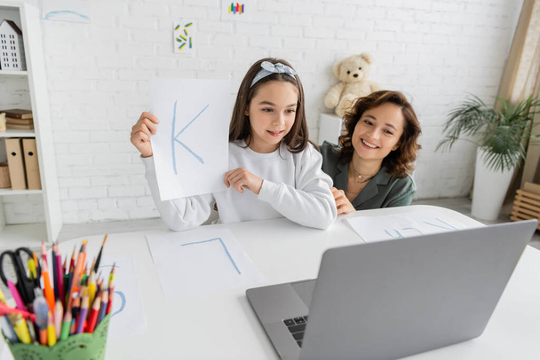 Веселый родитель смотрит на ноутбук рядом с дочерью с письмом, имеющие видео-урок логопедии дома  - Фото, изображение