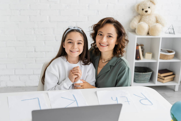 Cheerful woman and daughter looking at camera near papers with letters on desk  - Фото, зображення