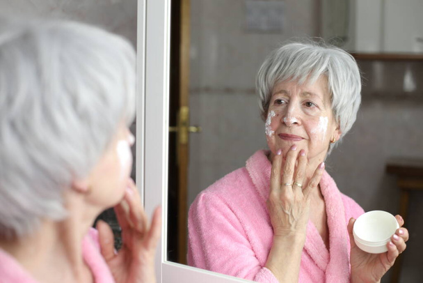 close-up retrato de mulher madura aplicando creme facial na frente do espelho no banheiro - Foto, Imagem