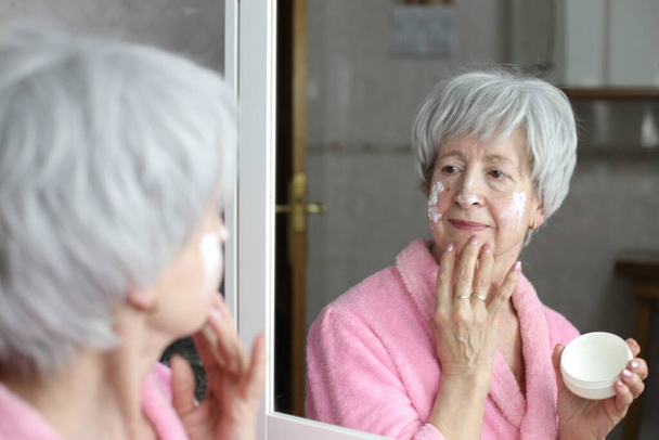 lähikuva muotokuva kypsä nainen soveltamalla kasvovoide peilin edessä kylpyhuoneessa - Valokuva, kuva