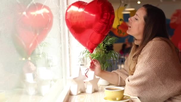 Día de San Valentín. Mujer joven bonita en suéter está sentado en la cafetería en la mesa por la ventana con la taza de capuchino y sonriendo soplando en el corazón rojo inflable. Momentos felices. Regalos y diversión. Imágenes de FullHD. - Metraje, vídeo