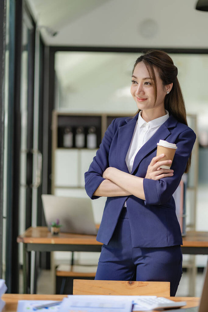 Güzel gülümseyen bir iş kadını elinde dizüstü bilgisayarla elinde kahve fincanıyla ayakta duruyor ve küçük işletmeleri yönetiyor. - Fotoğraf, Görsel