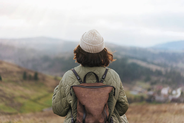 Widok z tyłu młodej turystki w plecaku i czapce stojącej na krawędzi klifu i cieszącej się krajobrazem, zachodem słońca, górami i chmurami. - Zdjęcie, obraz