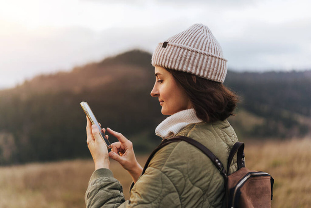 Happy smiling female traveller using cell telephone with online map and texting to friends, blurred forest and mountains in the background, concept of using technologies in nature - 写真・画像