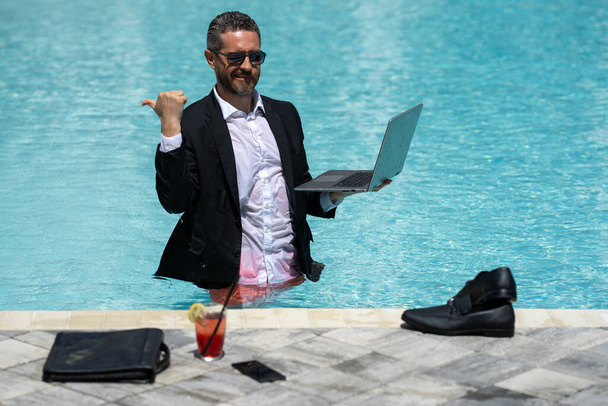 Leisure weekend and remote freelance work. Crazy comic business. Funny businessman in suit with laptop in swimming pool water. Remote summer work. Businessman relaxing in a swimming pool in a suit - Photo, image