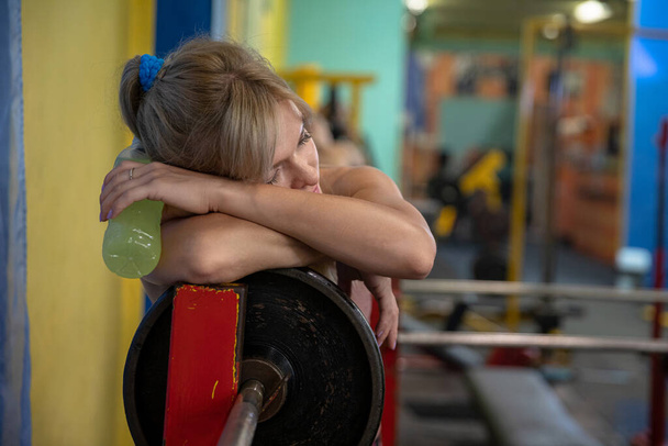 A young woman is leaning with her hands on an exercise barbell. She rests her head on her hands and has her eyes closed. In her hand she holds a bottle of yellow isotonic drink. - 写真・画像