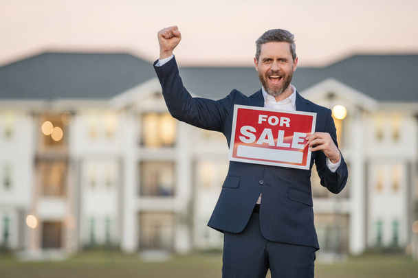 Portrait of american real estate agent standing outdoors at house with board for sale. Real estate broker with sign for sale. Finance and banking concept. Home purchase mortgage concept - Foto, Imagem