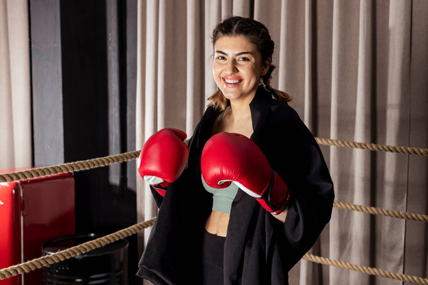 Una hermosa chica sonriente con guantes de boxeo rojos se para en una postura de lucha y mira a la cámara en un ring de boxeo. - Foto, imagen