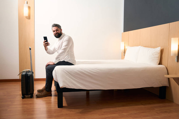 Joyful man with mobile phone in hand sitting on edge of bed in hotel room - Fotografie, Obrázek