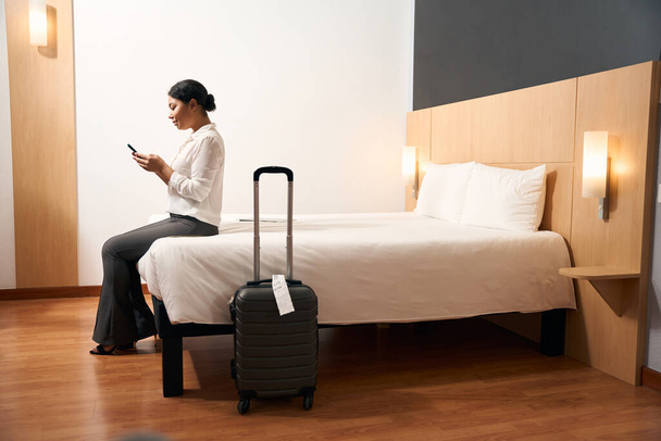 Zijaanzicht van geconcentreerde vrouwelijke toerist met mobiele telefoon in handen zittend op rand van bed in hotelkamer - Foto, afbeelding