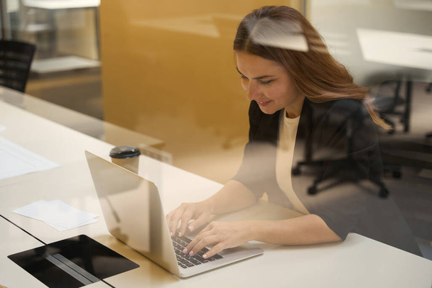 Femme souriante assise à table et tapant sur le clavier de l'ordinateur dans le bureau - Photo, image