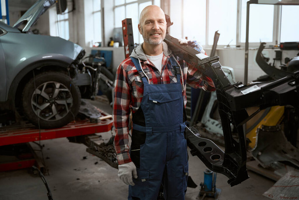 Strong master holds a part of the car body on his shoulder, a man in work overalls - Photo, image