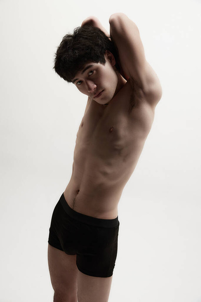 Portrait of young man posing shirtless in black underwear against grey studio background. Fit body, attentive look. Concept of male body aesthetics, style, fashion, health, wellness - 写真・画像