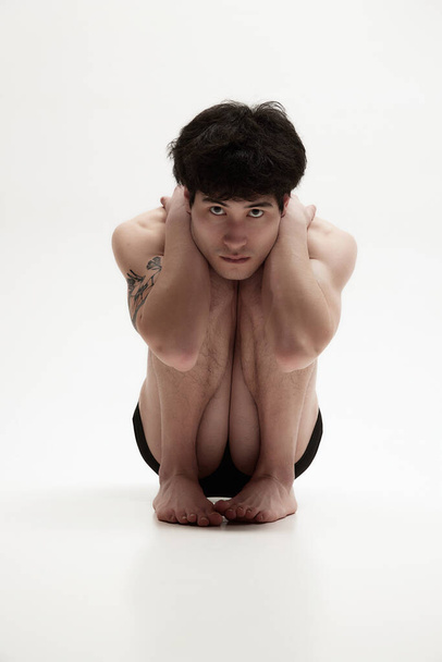 Inner fears. Portrait of young serious guysittign shirtless, in underwear on floor and holding himself with hands against white background. Concept of male body aesthetics, style, health, mens beauty - Foto, imagen