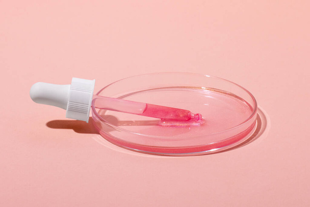 De cosmetische pipet ligt in een petrischaal met rozengoud. Of een roze vloeistof met glitter. Laboratorium onderzoek van cosmetica, gel, geneeskunde. Scheikunde - Foto, afbeelding
