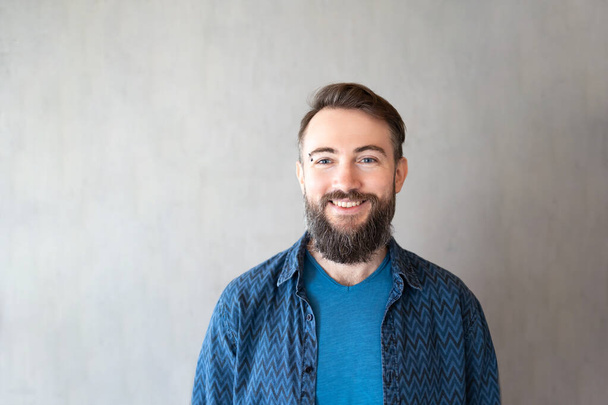 Close-up portrait of bearded biracial handsome young man with eyebrow piercing smiling against grey wall, copy space - Photo, Image