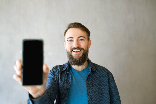 Advertentie voor mobiele apps. Knappe Spannende Man toont Pointing at Empty Smartphone Screen poseren over grijze achtergrond, glimlachen om camera. Kijk hier eens, mobiele telefoon weergave Mock Up - Foto, afbeelding